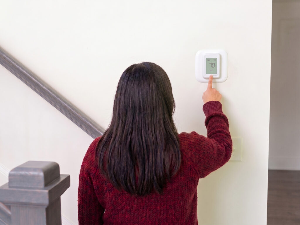 a homeowner adjusts her thermostat, currently set to 70 degrees, in the hallway near her staircase