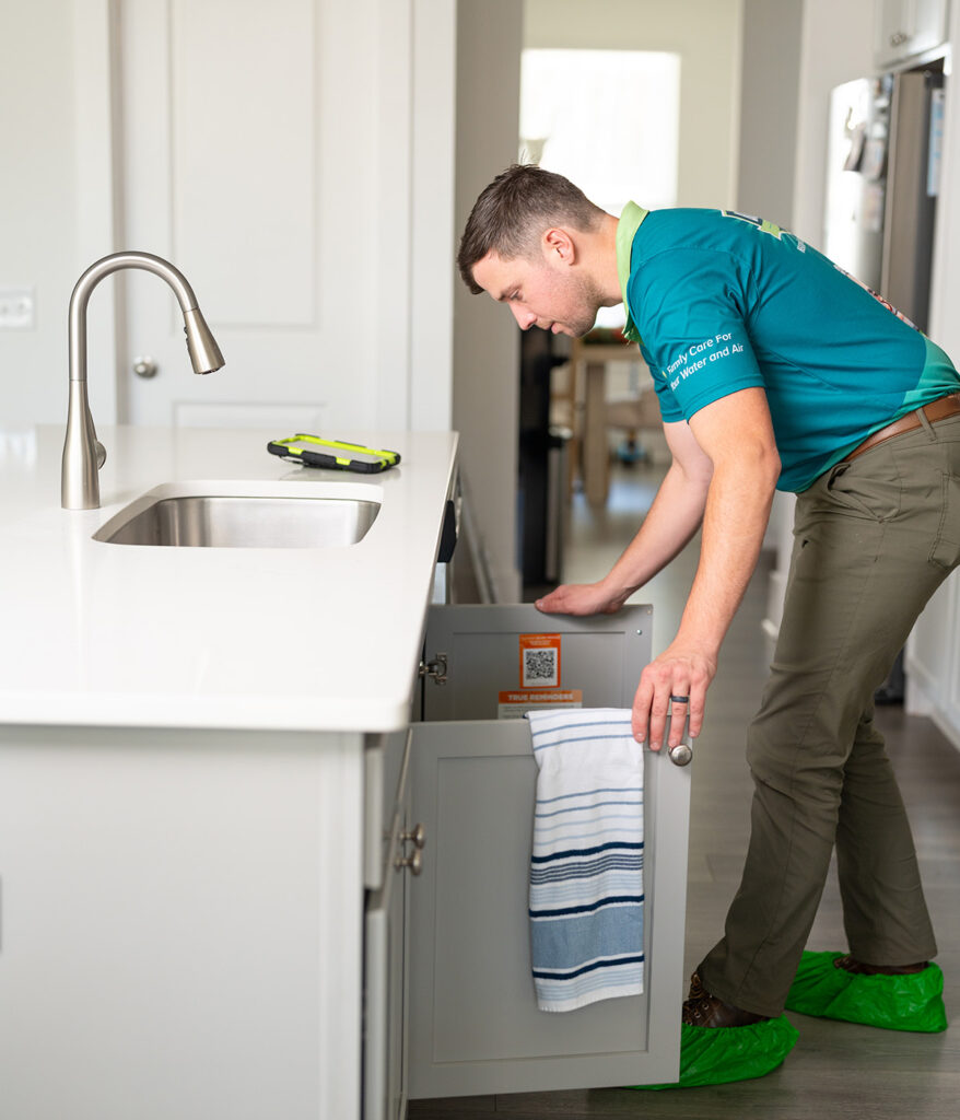 lanton & Sons plumber wearing shoe covers opening under-sink cabinet to inspect plumbing in a modern white kitchen